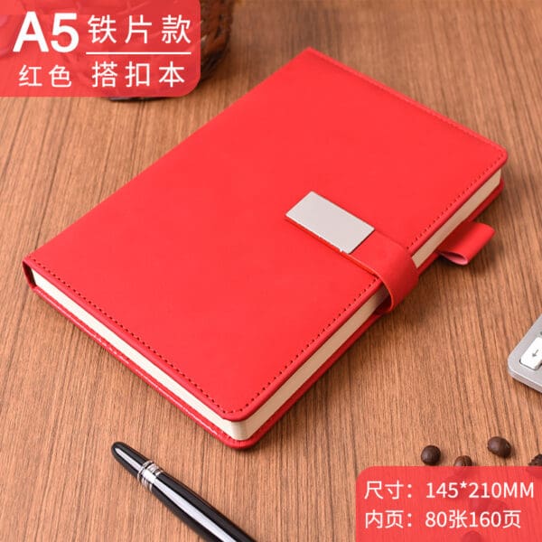 Red A5 notebook with metallic clasp, black pen, and coffee beans on a wooden table.